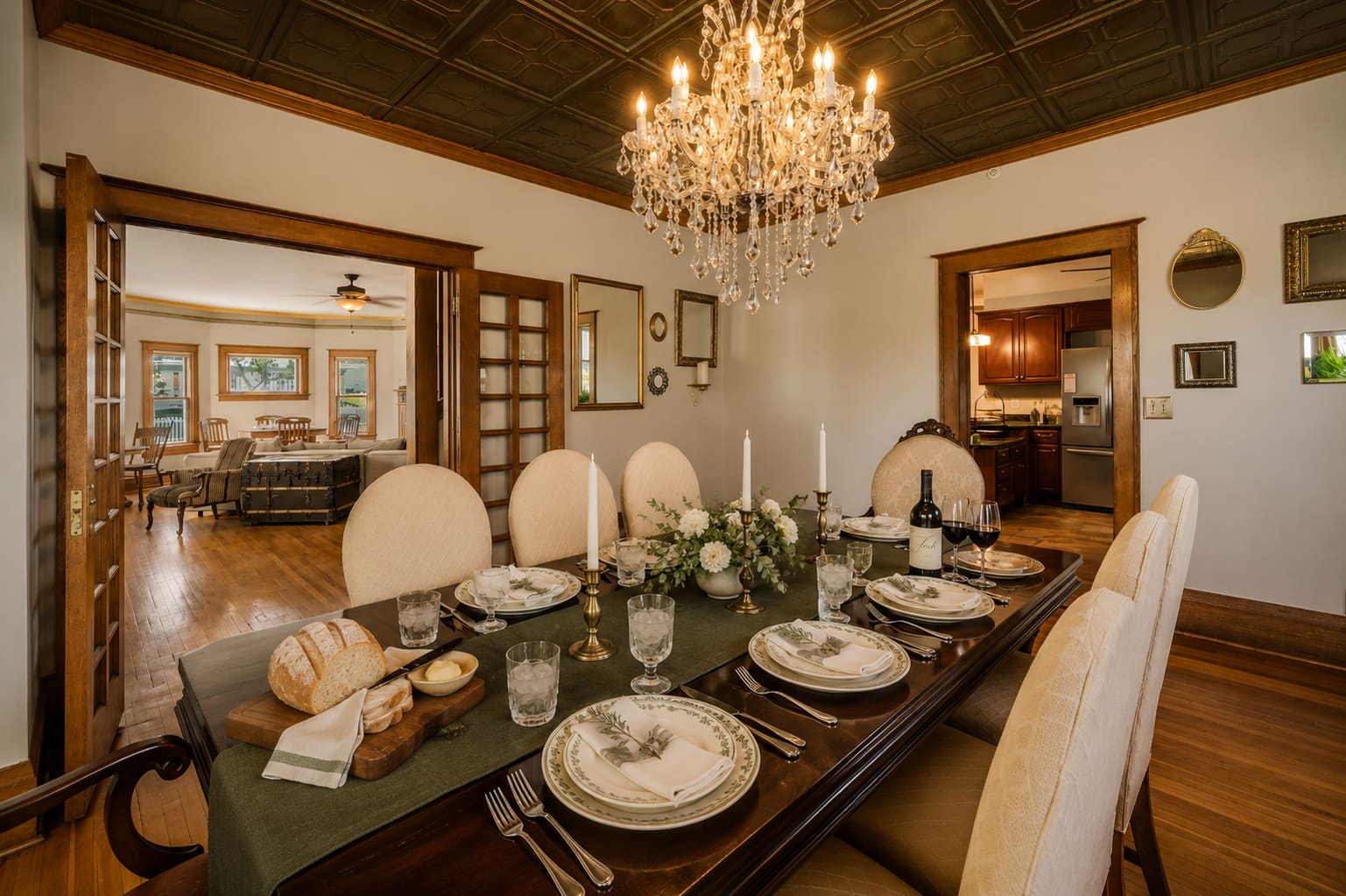 The Dining Room at Gratitude Sheridan — long mahogany table set for twelve under a crystal chandelier, candles lit, bread board and wine, the Parlor visible through open pocket doors.